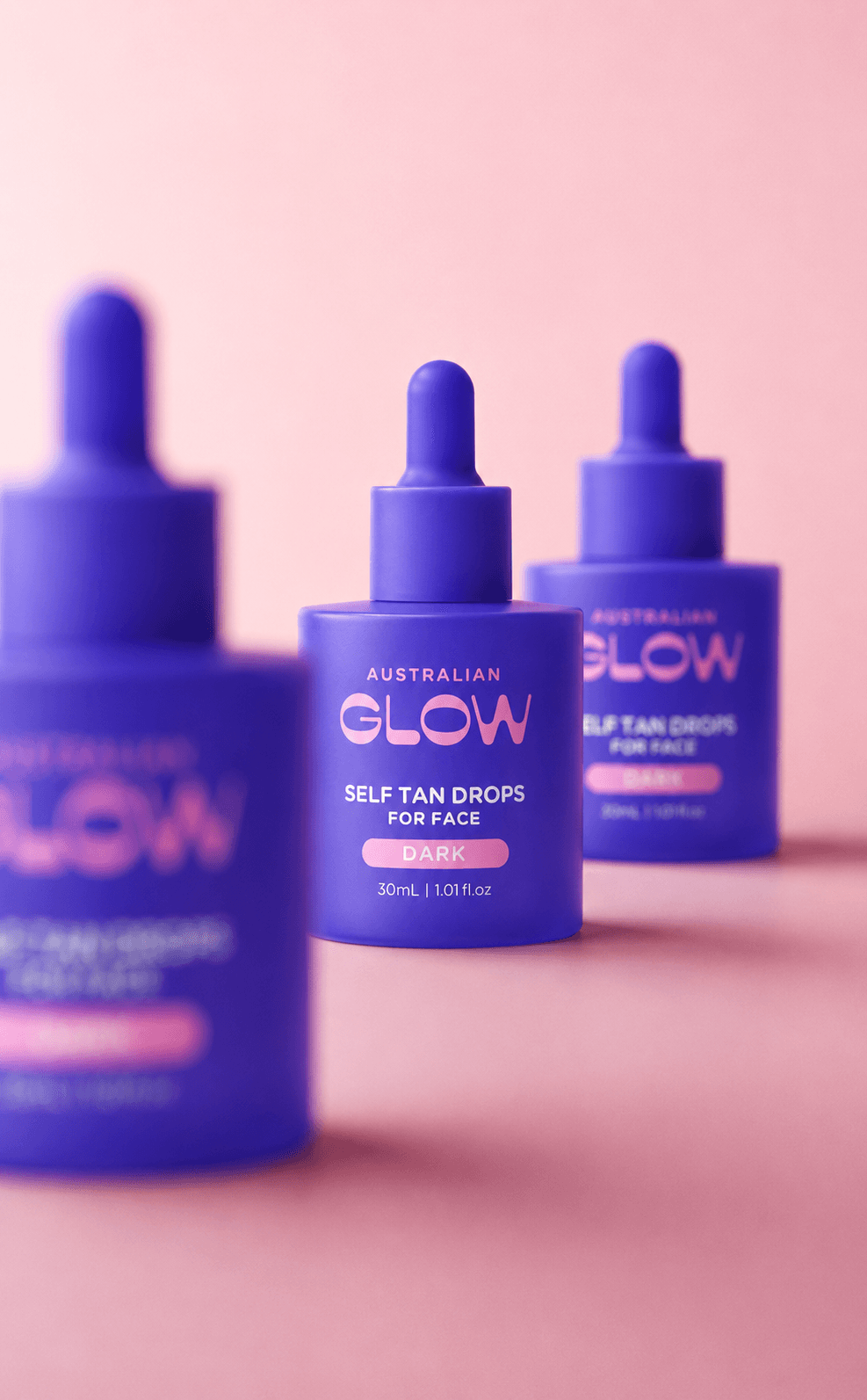 Face Tan Drops Dark — three bottles arranged in a row on a pale-pink studio backdrop